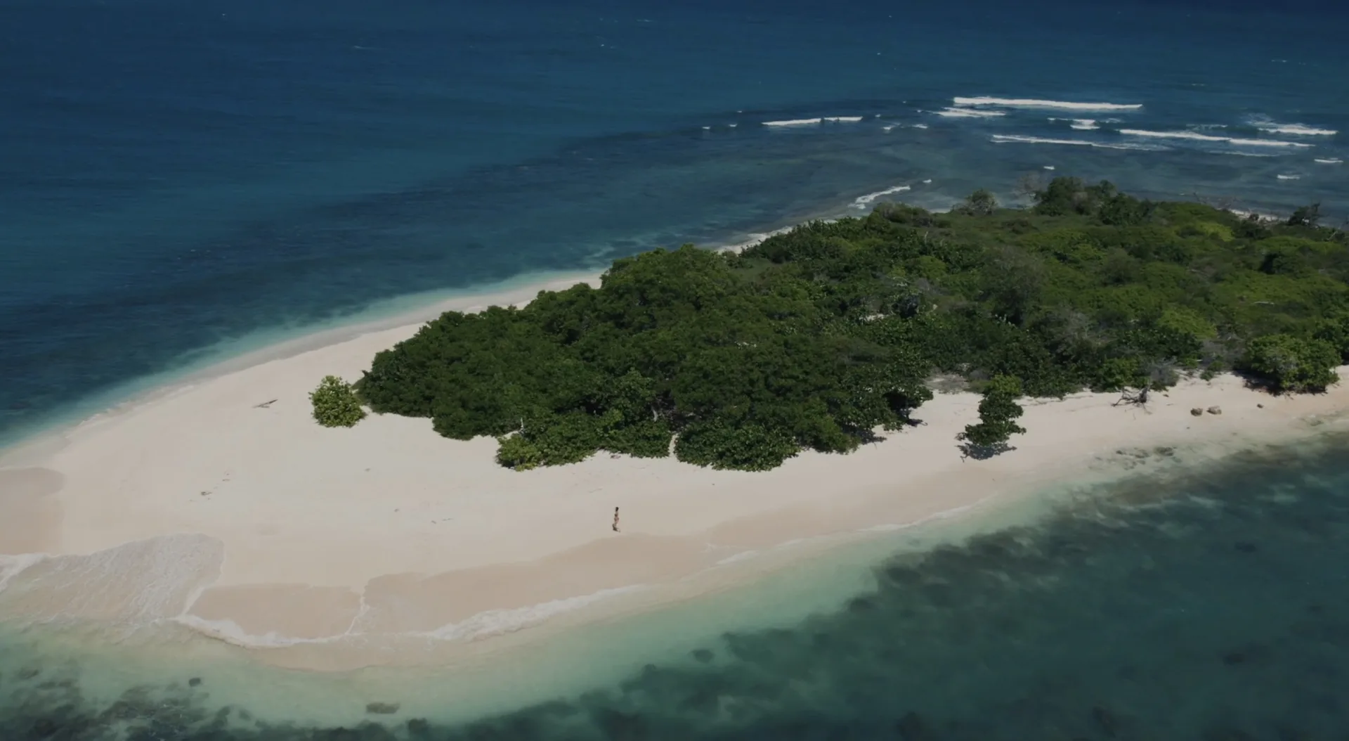 Aerial view of a small tropical island with dense green trees, white sandy beach, and clear turquoise water.