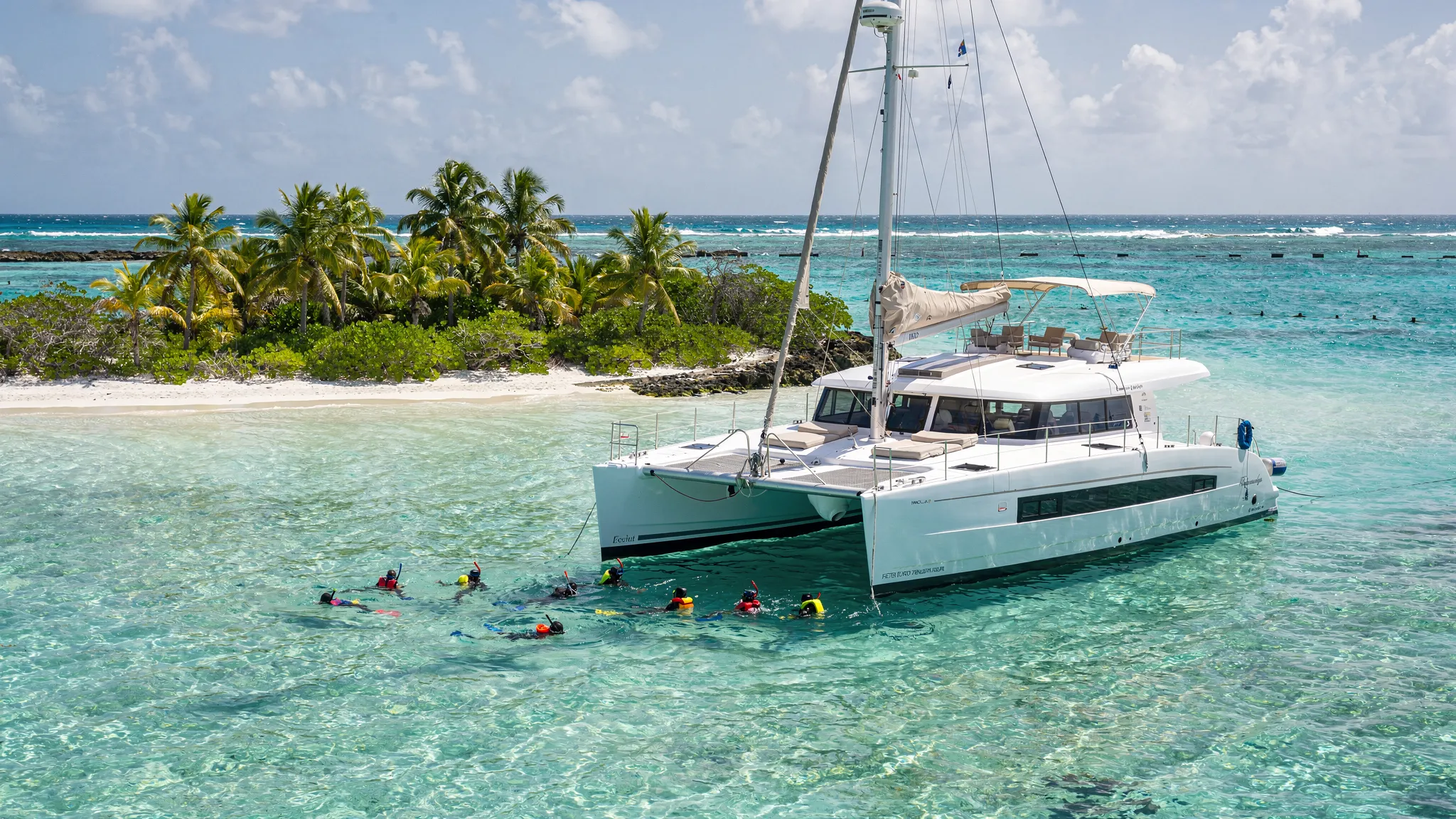 A luxury crewed catamaran anchored in calm turquoise water near a palm-lined Belize caye, with the Belize Barrier Reef visible as a darker line in the distance, and guests snorkeling near the boat in clear, shallow water.