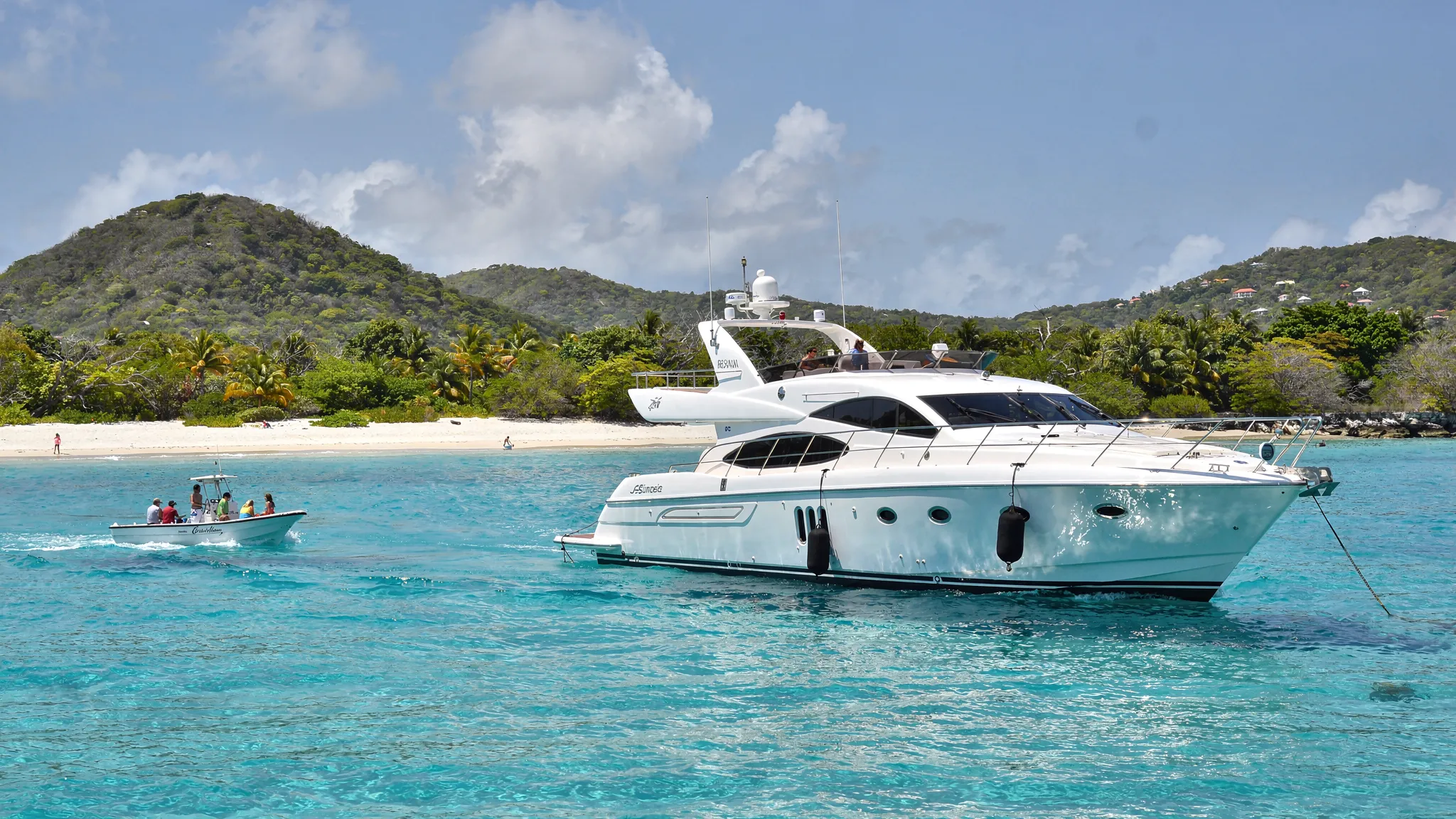A luxury yacht anchored in clear turquoise water near a white-sand beach in the US Virgin Islands, with a tender heading toward shore and green hills in the background.