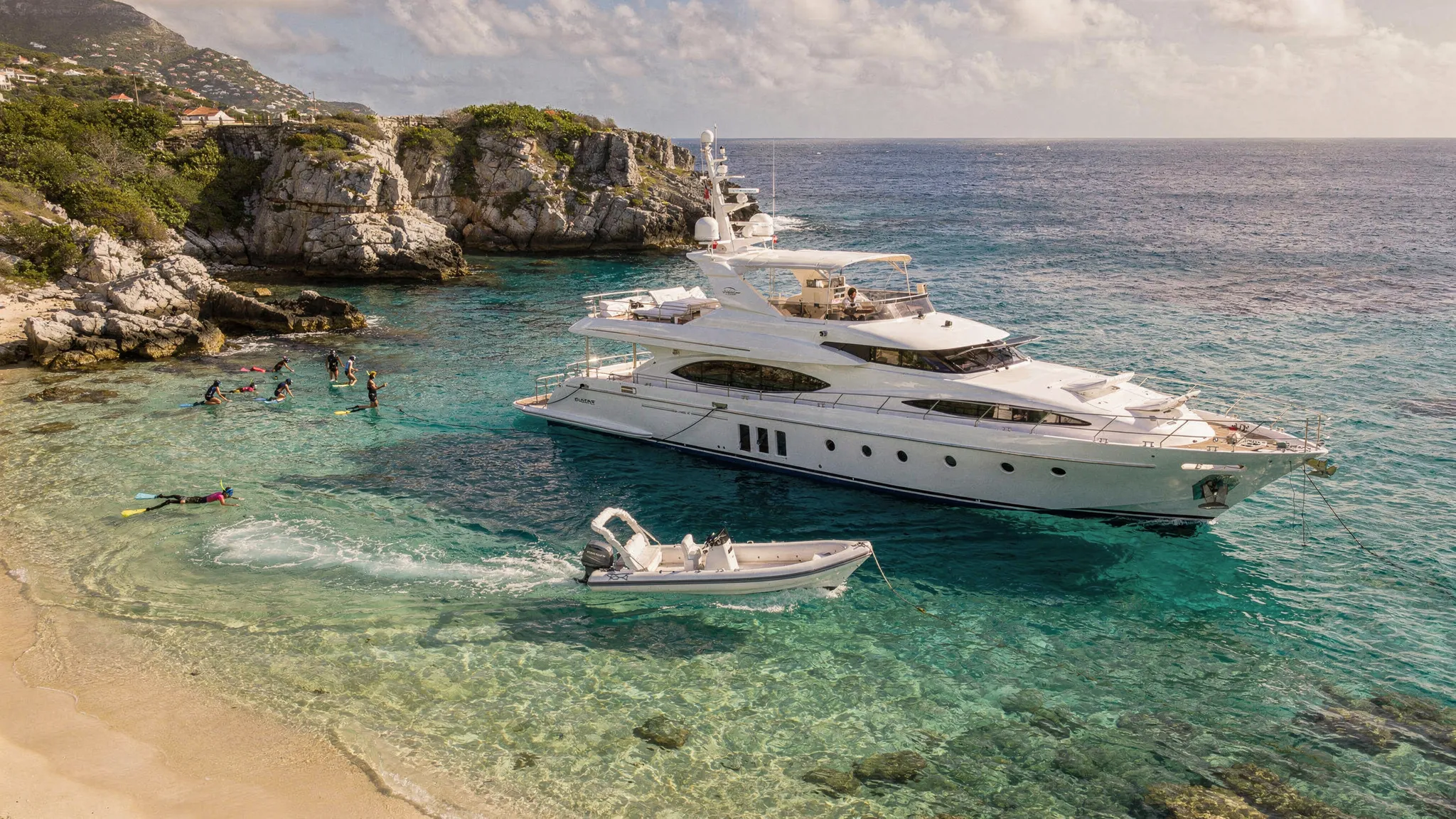 A luxury yacht anchored off a quiet St. Barts bay with turquoise water, a tender heading to shore, and guests snorkeling near a rocky edge in the background.