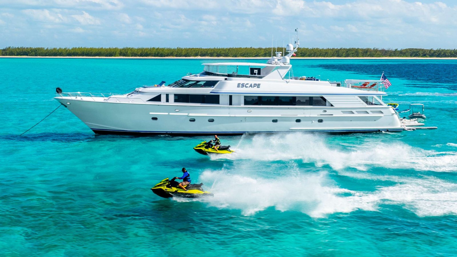 White luxury yacht named 'Escape' sails on bright turquoise water near a sandy shore, with two people riding yellow jet skis in front and spray behind them.
