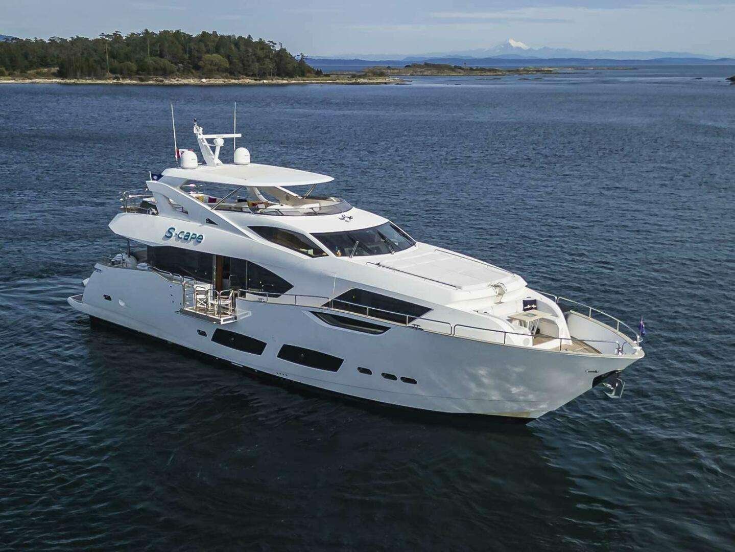 White luxury motor yacht on calm blue water with a forested shoreline in the distance and a clear sky above.