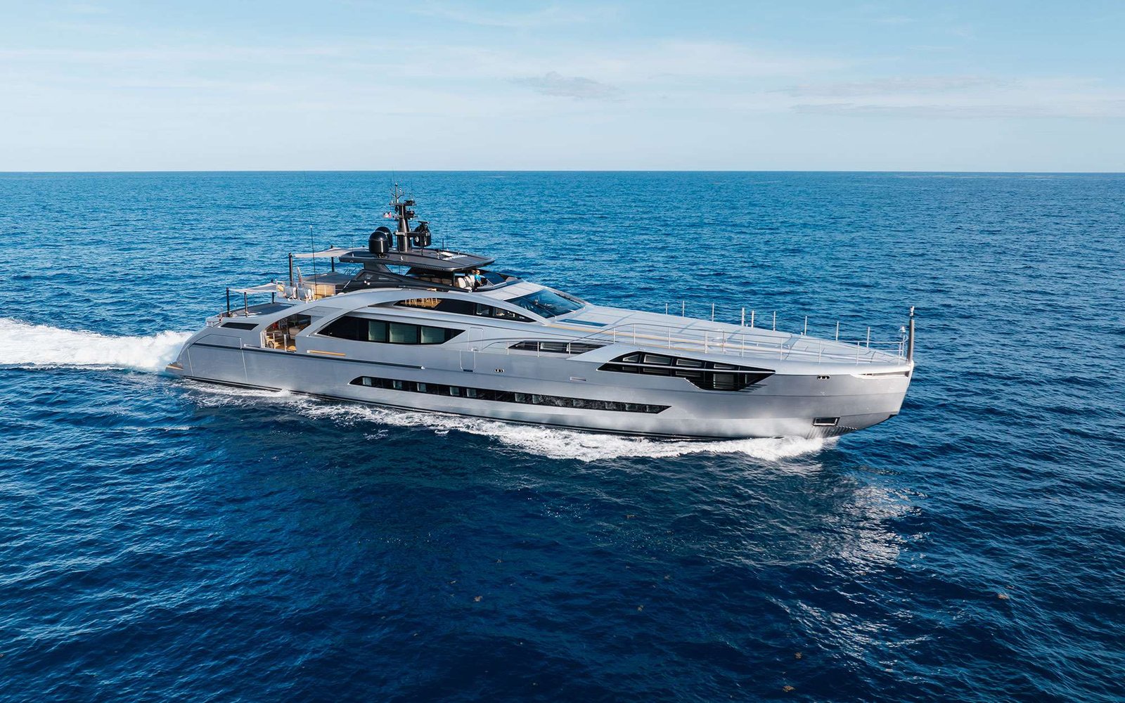 Sleek silver luxury yacht cruising on deep blue ocean under a clear sky, leaving a white wake behind it.