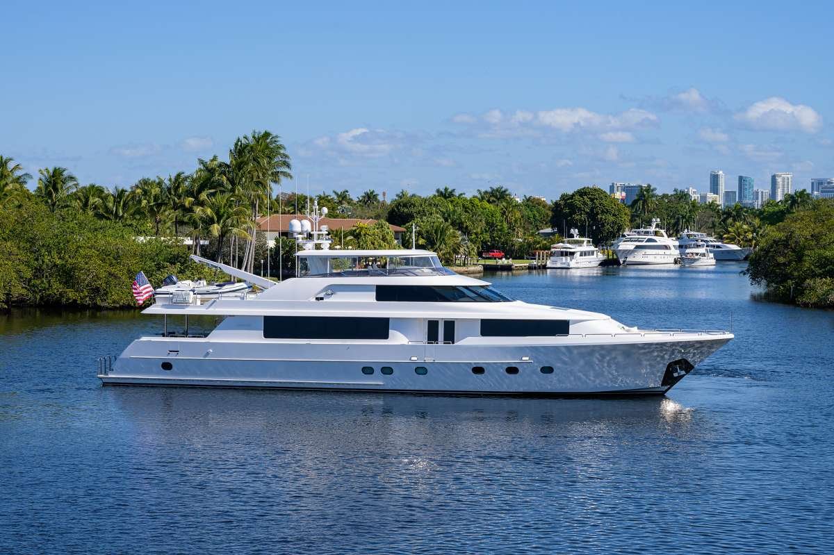 White luxury yacht on calm blue water with palm-lined shore and other boats in the background