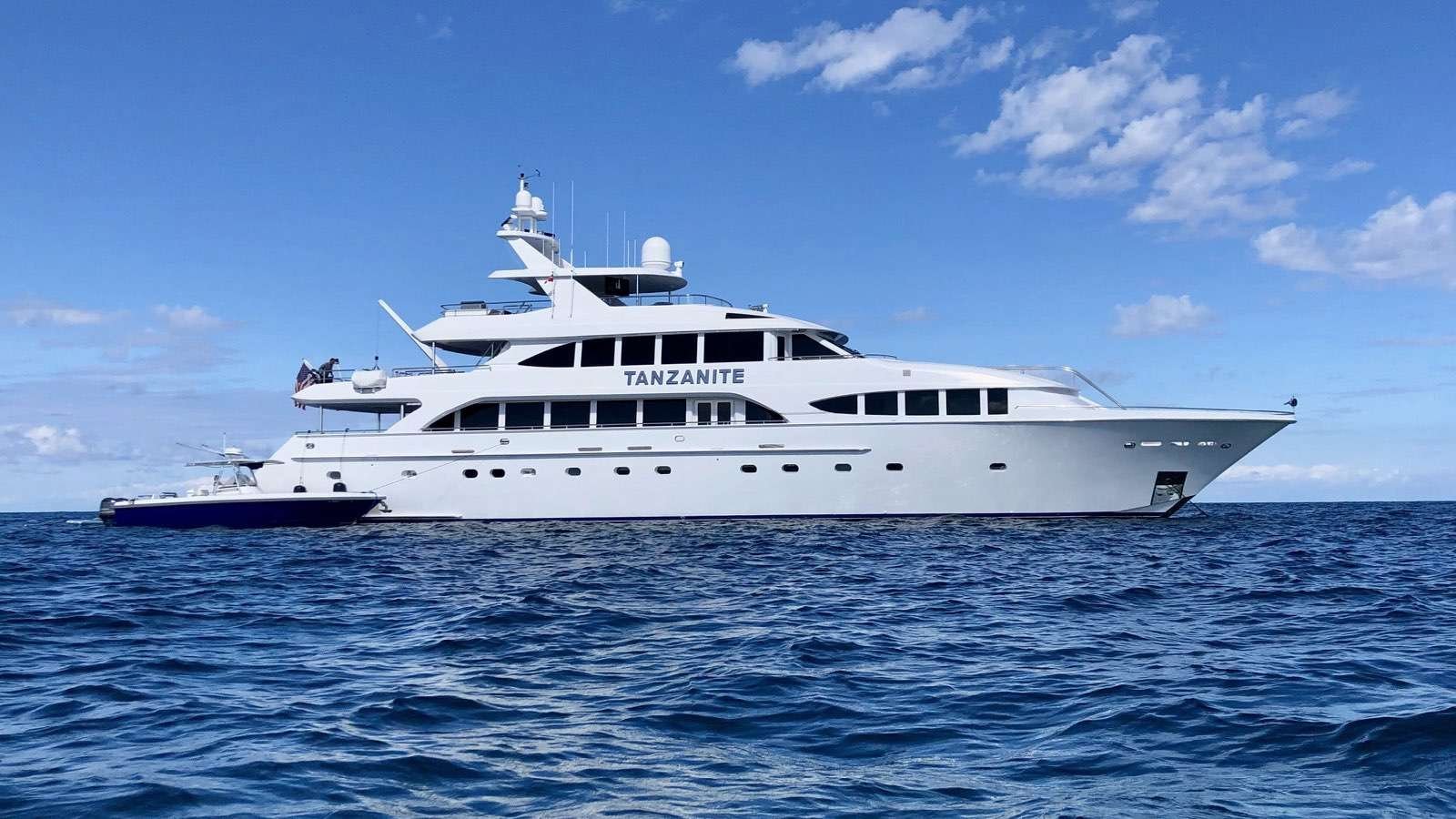 Large white luxury yacht named Tanzanite cruising on deep blue ocean, with a smaller boat alongside.