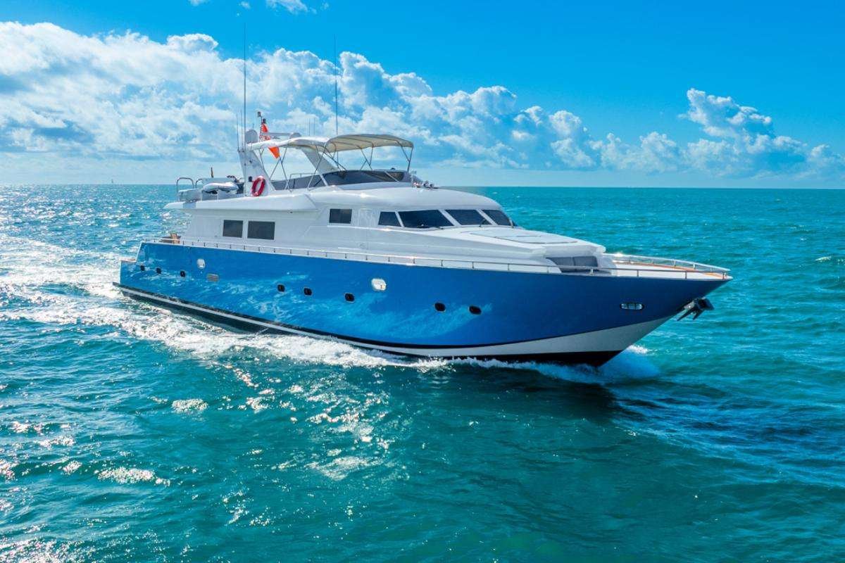 White-and-blue motor yacht cruising on clear turquoise water under a bright blue sky with fluffy clouds, creating white wake.