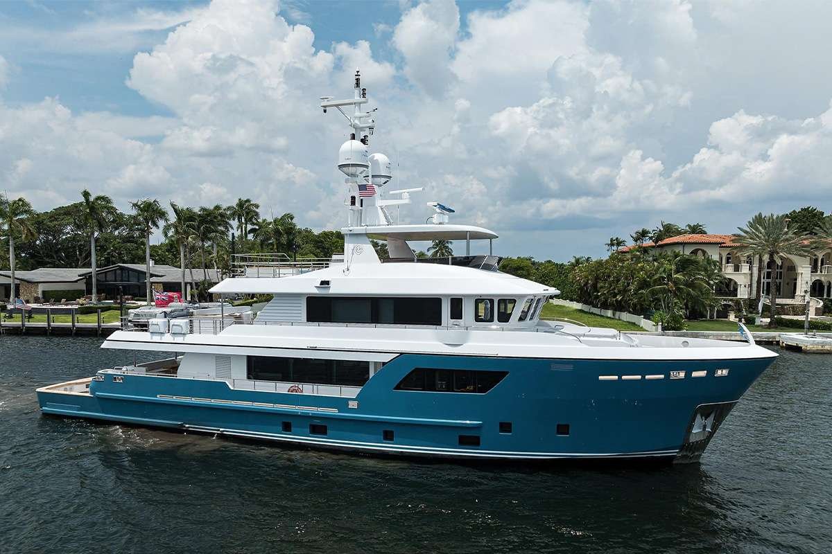 Blue and white luxury motor yacht on calm water with a palm-lined waterfront and resort homes in the background.