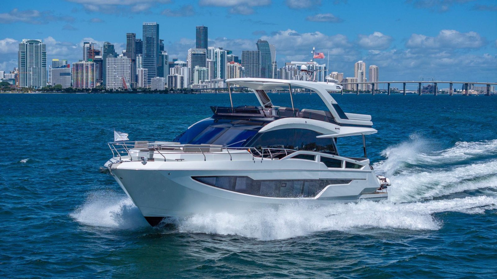 White luxury motor yacht cruises on blue water with a city skyline in the background, flag waving atop the boat.