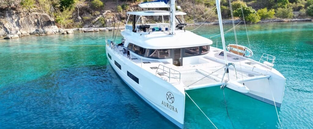 White catamaran named 'Aurora' anchored in clear turquoise water near rocky shoreline, with a small dinghy on deck and a sunny sky.