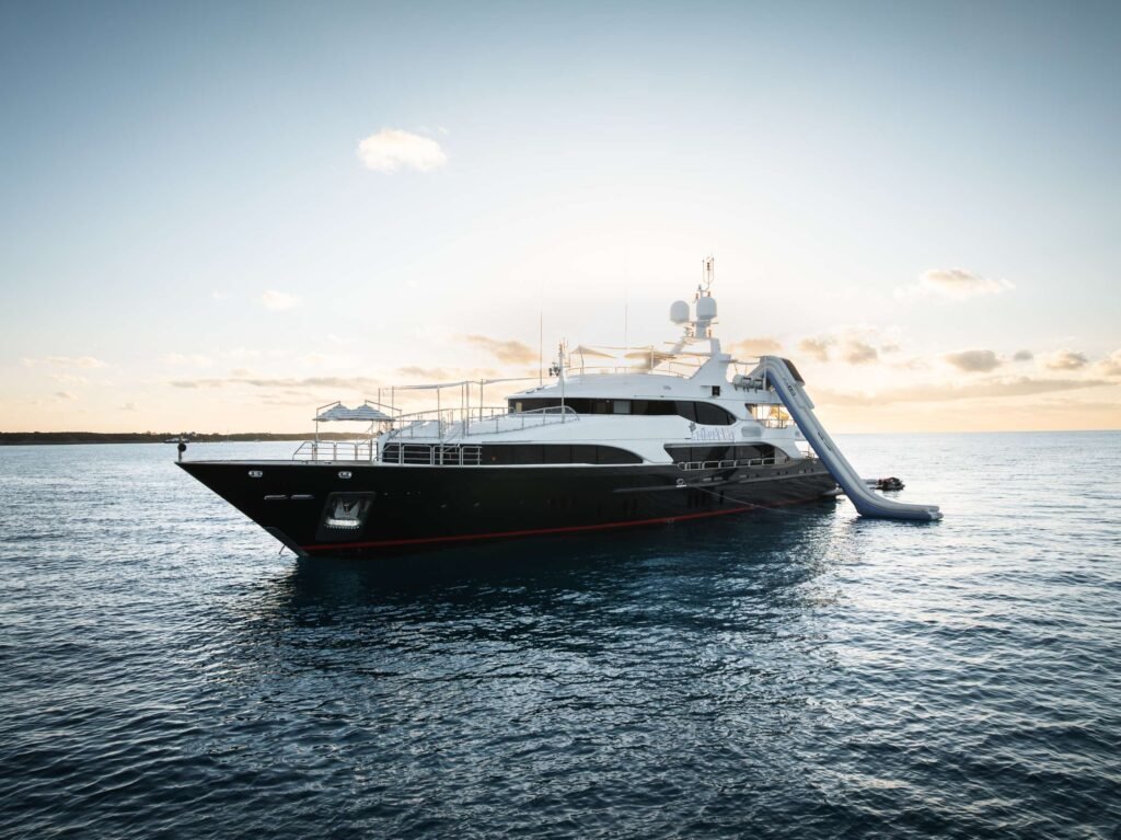 Large white luxury yacht on calm water at sunset with a clear sky and horizon in the background, a gangway extending from the side to a dock edge.