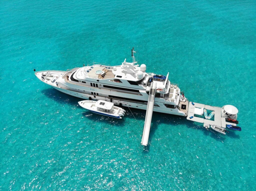 Aerial view of a white luxury yacht moored at a dock in clear turquoise water, with a small boat nearby and jet skis on the platform.