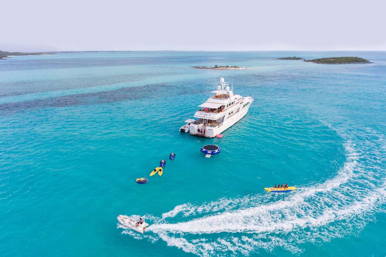 A white luxury yacht sails in clear turquoise water near small tropical islands, with people on deck and jetskis nearby.