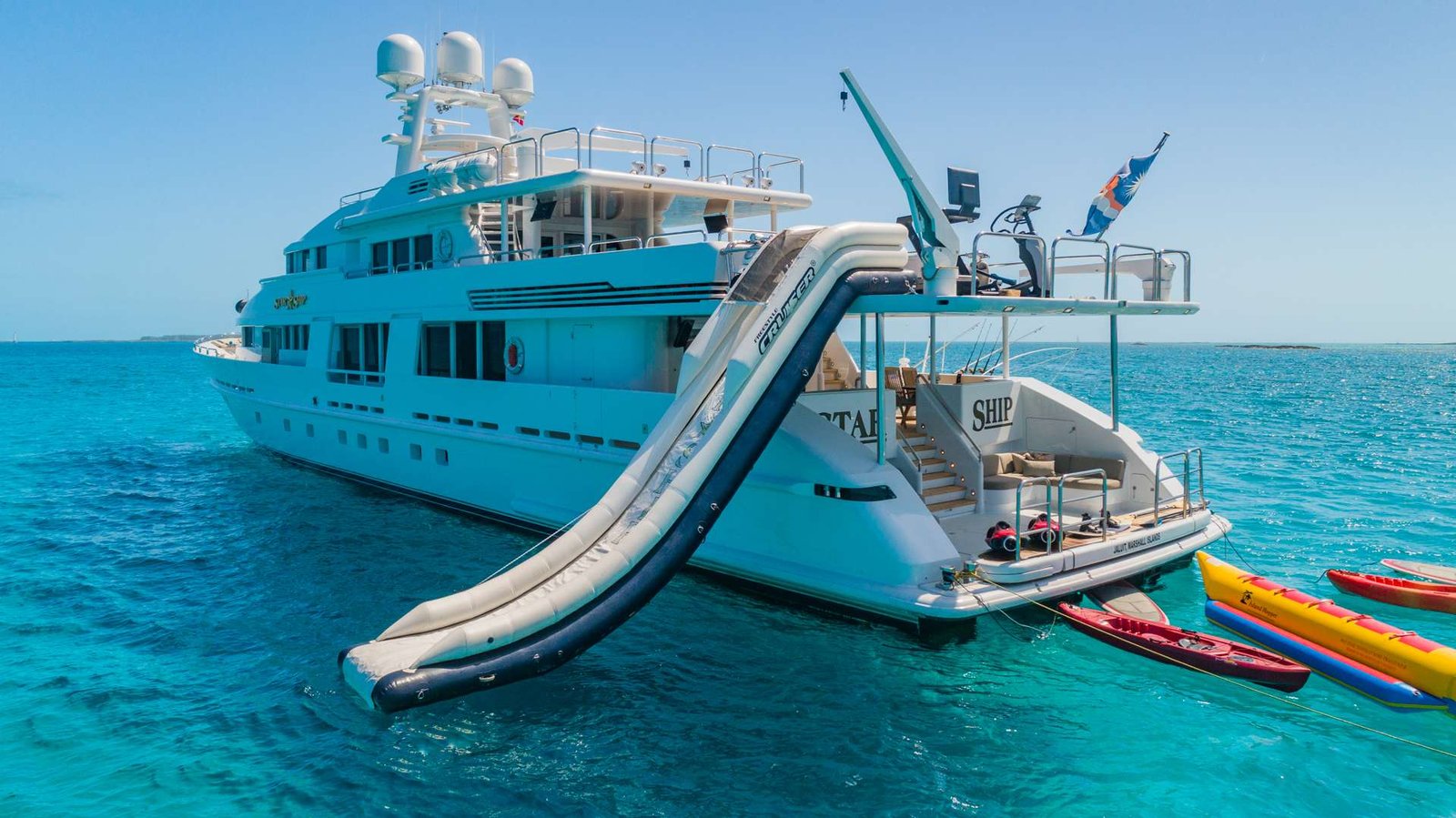 Large white luxury yacht with inflatable water slides attached, anchored in turquoise sea with colorful kayaks nearby.