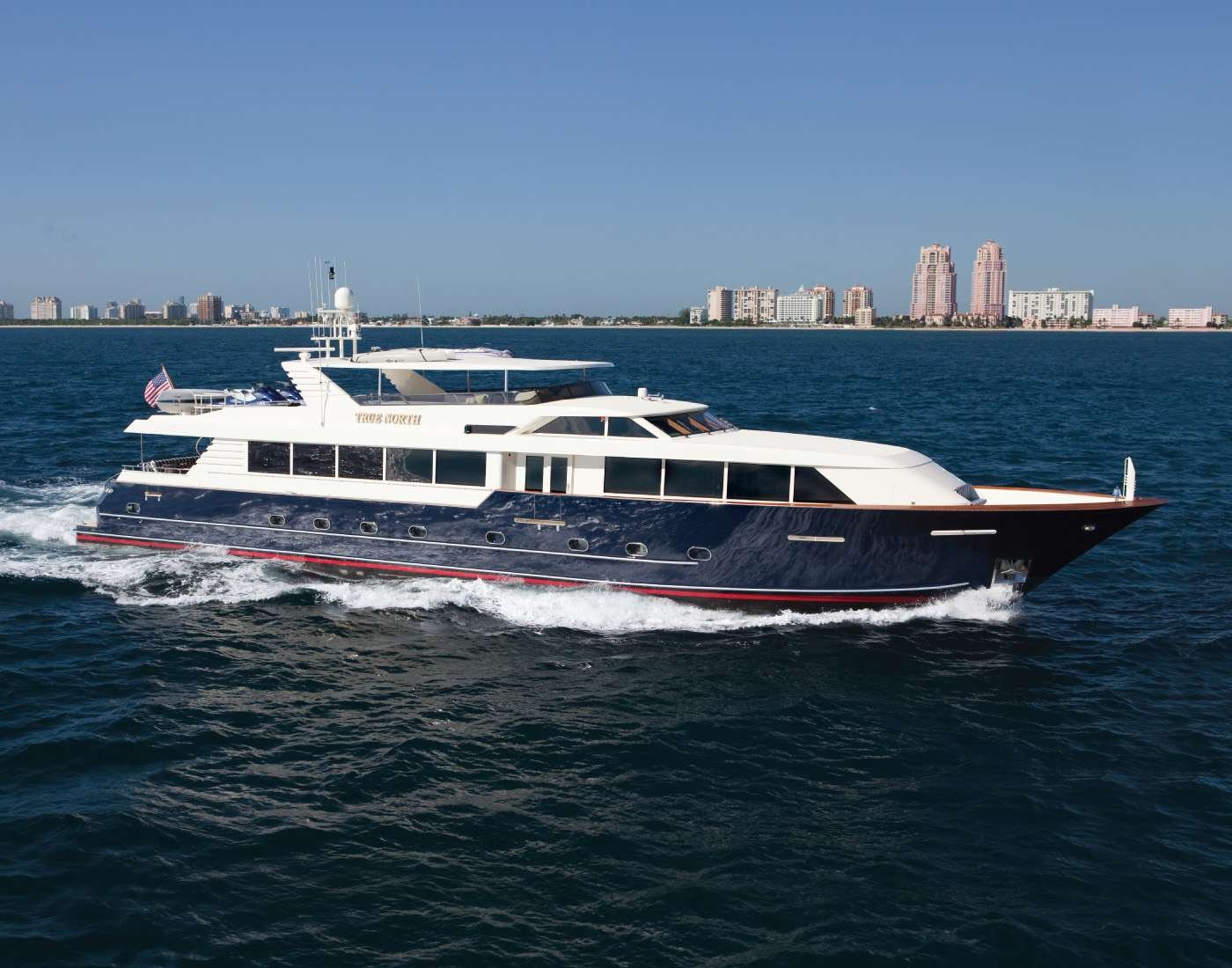 Large white and navy luxury motor yacht cruising on blue ocean with a city skyline in the distance on a sunny day.