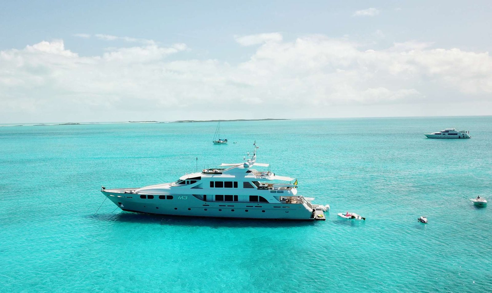 Large white luxury yacht anchored in clear turquoise water, with smaller boats nearby and a distant horizon under a blue sky.