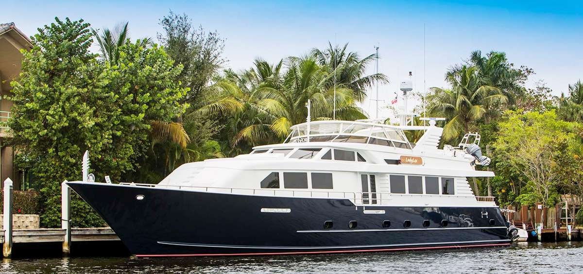 Large white and navy motor yacht docked at a tropical waterfront with palm trees and a wooden seawall nearby.