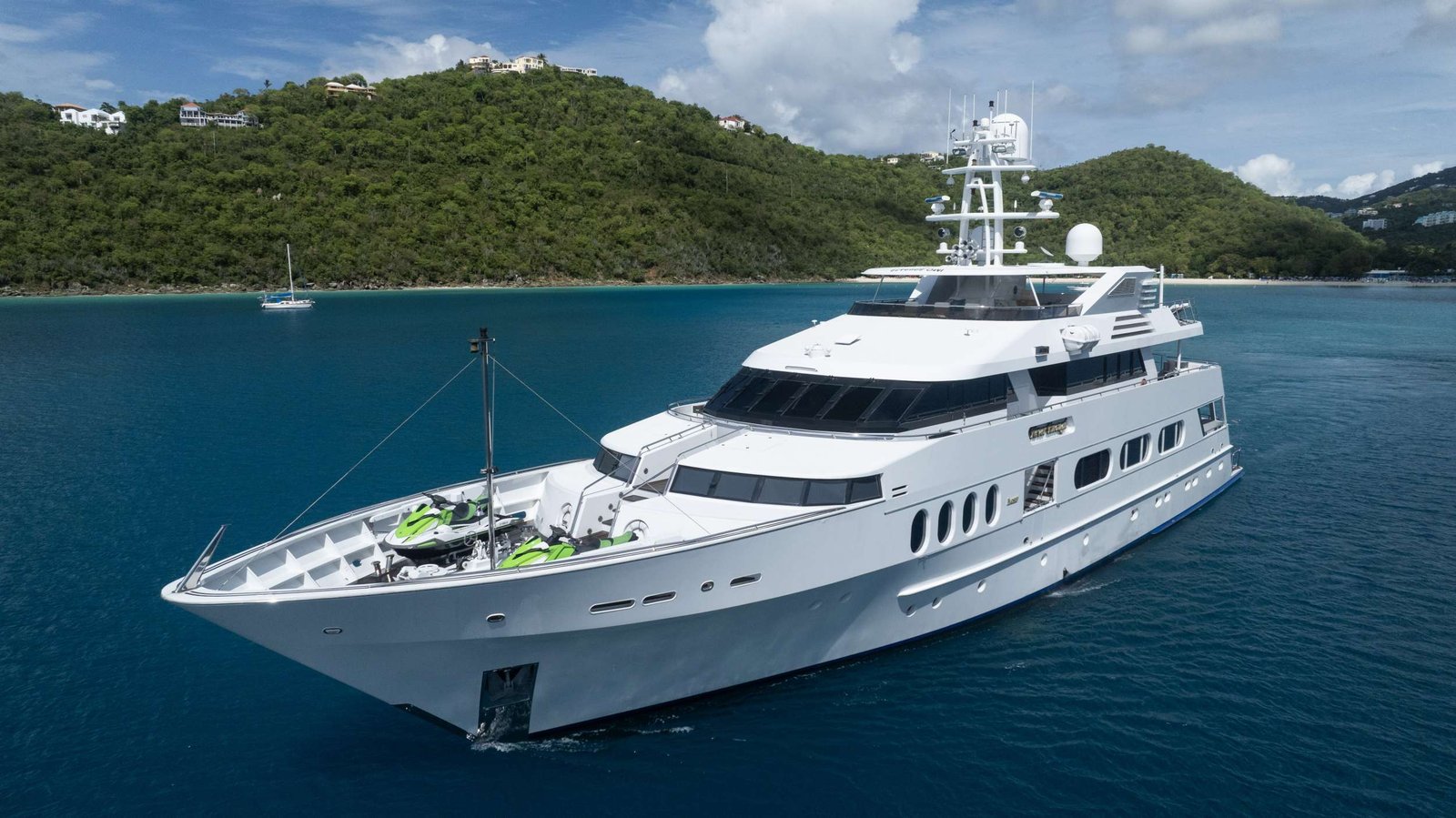 Large white luxury yacht in turquoise water near a green, hilly coastline with houses on the hillsides in the background.