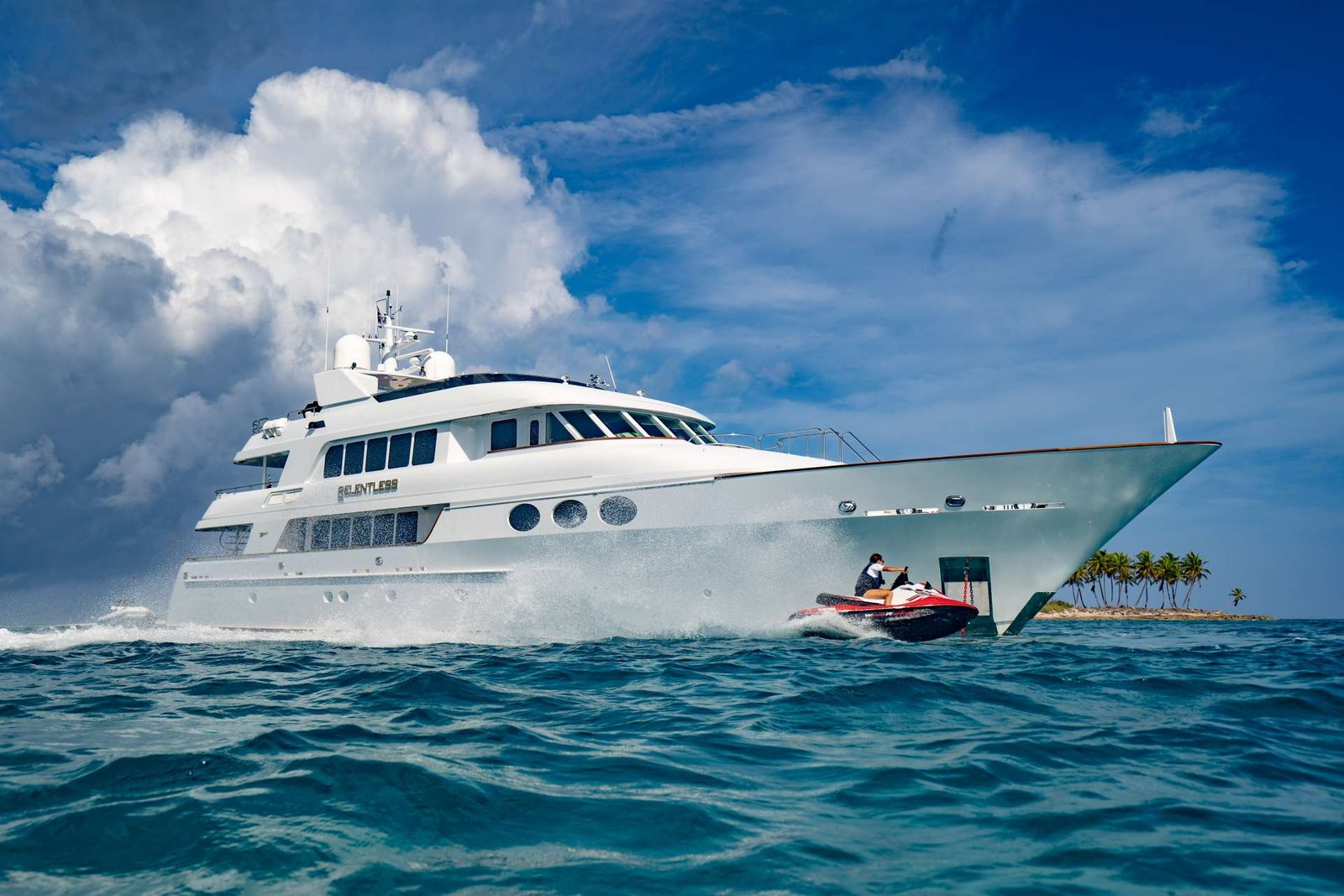 White luxury yacht cruising on blue water with a person riding a red jet ski nearby and a palm‑fringed island in the distance