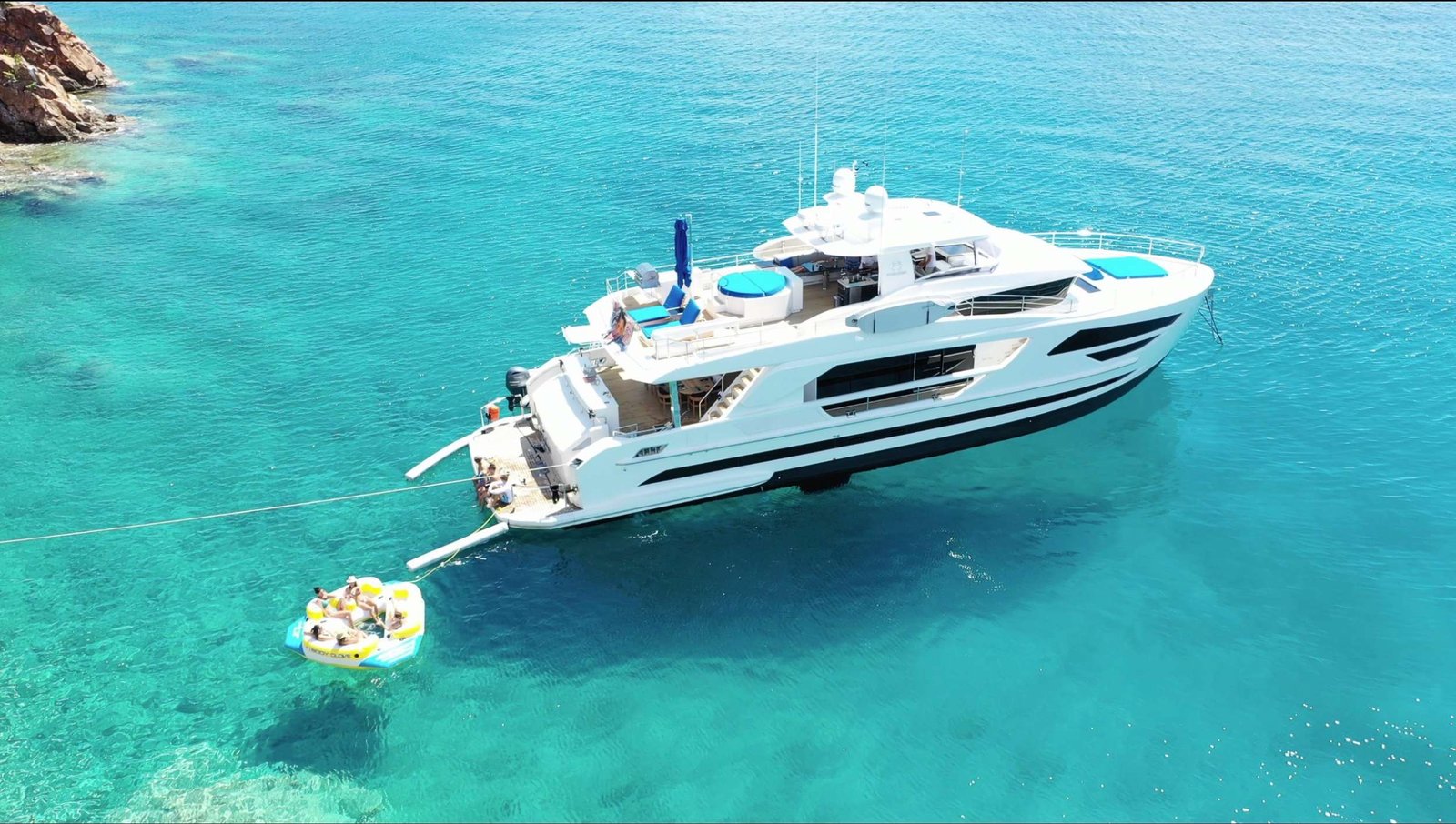 A white luxury yacht anchored in clear turquoise water with a small inflatable raft nearby carrying people outdoors on a sunny day in the sea.