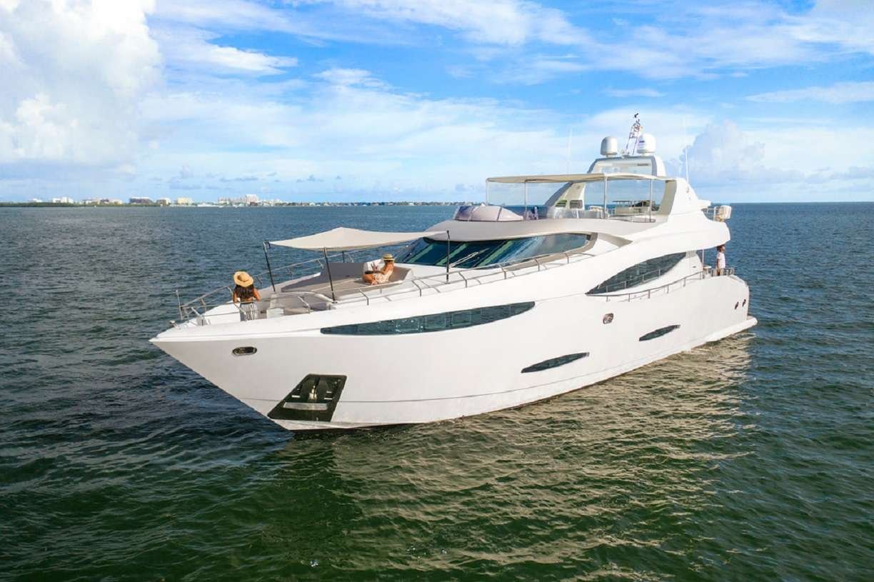 White luxury yacht cruising on calm sea under a blue sky with two people lounging on the deck's shaded area