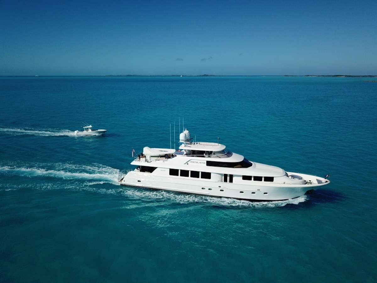 A large white luxury motor yacht cruises on bright blue ocean water, with a smaller tender boat trailing in its wake.