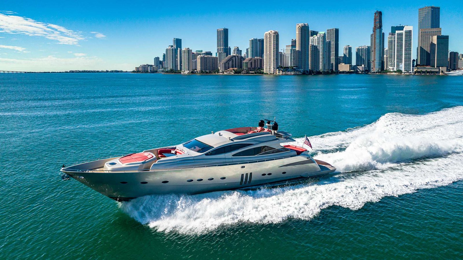 White luxury motor yacht cruises on blue-green water with a city skyline in the distance, wake trailing behind.
