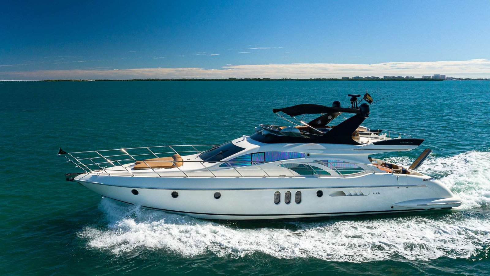 White motor yacht speeding across turquoise waters with wake behind it and a clear blue sky above, seen from the side of the hull.