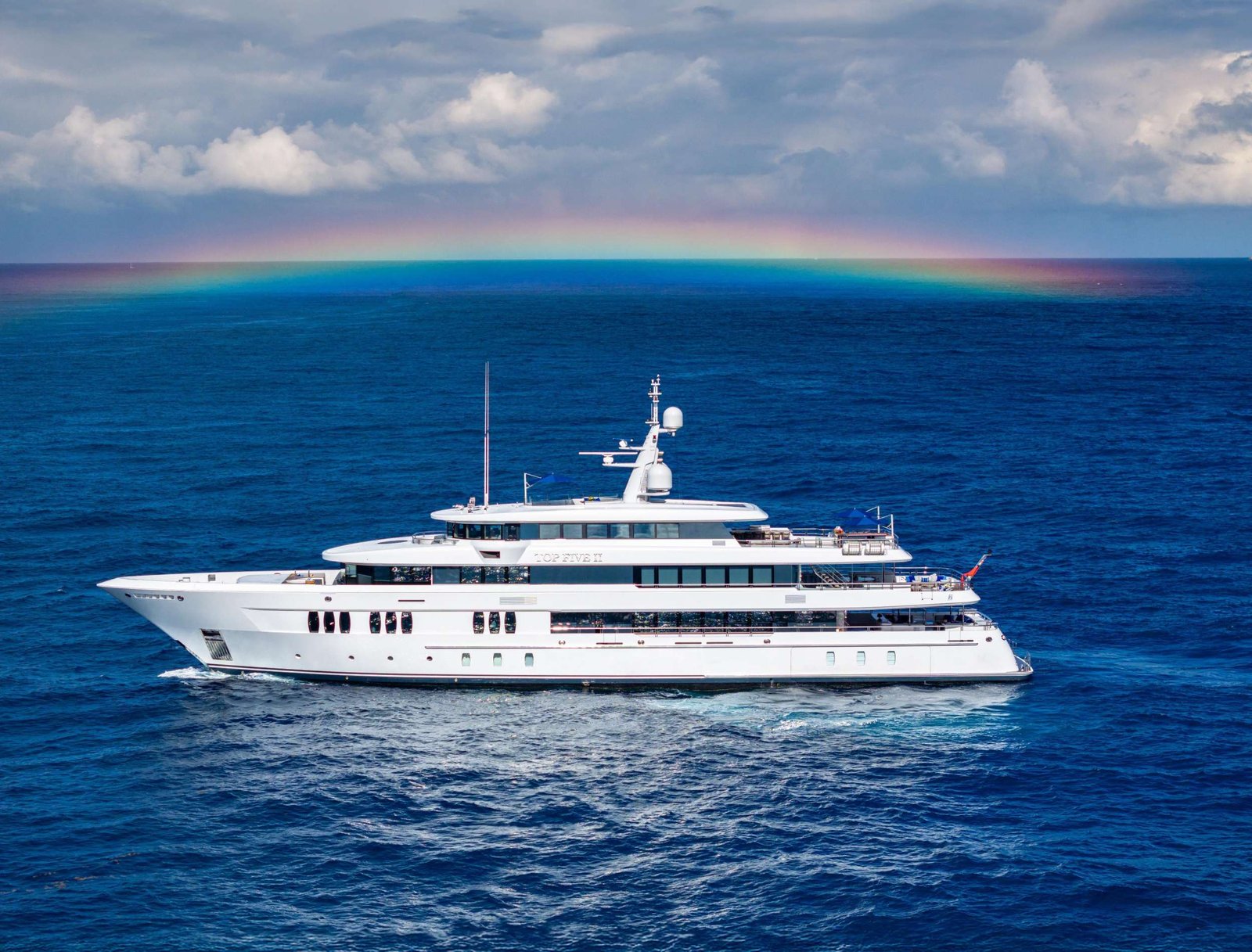 White luxury yacht sailing on deep blue ocean under a pastel rainbow and cloudy sky.