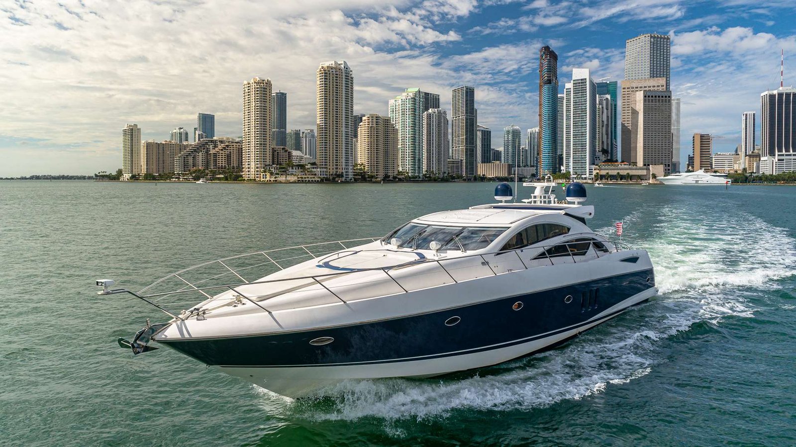 White luxury yacht sails across calm harbor with a high-rise city skyline in the background, under a partly cloudy blue sky.