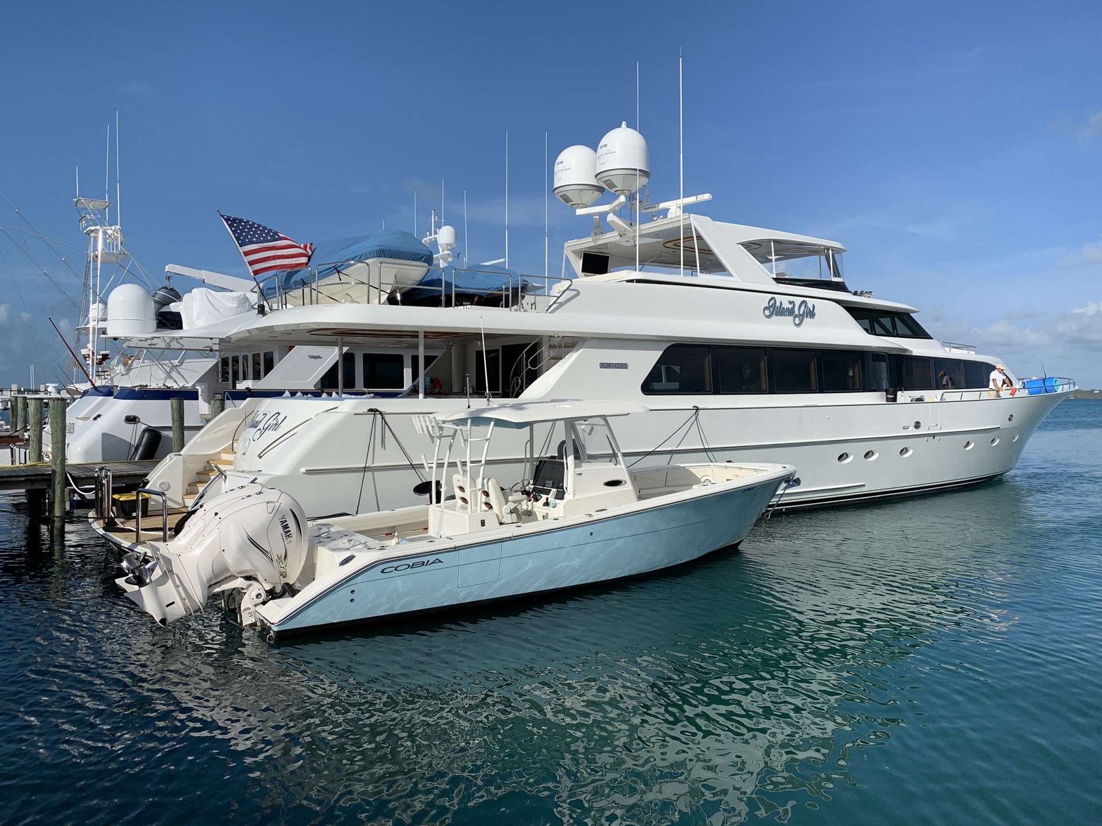 Two white luxury motor yachts docked at a marina under a clear blue sky, with an American flag fluttering between them.