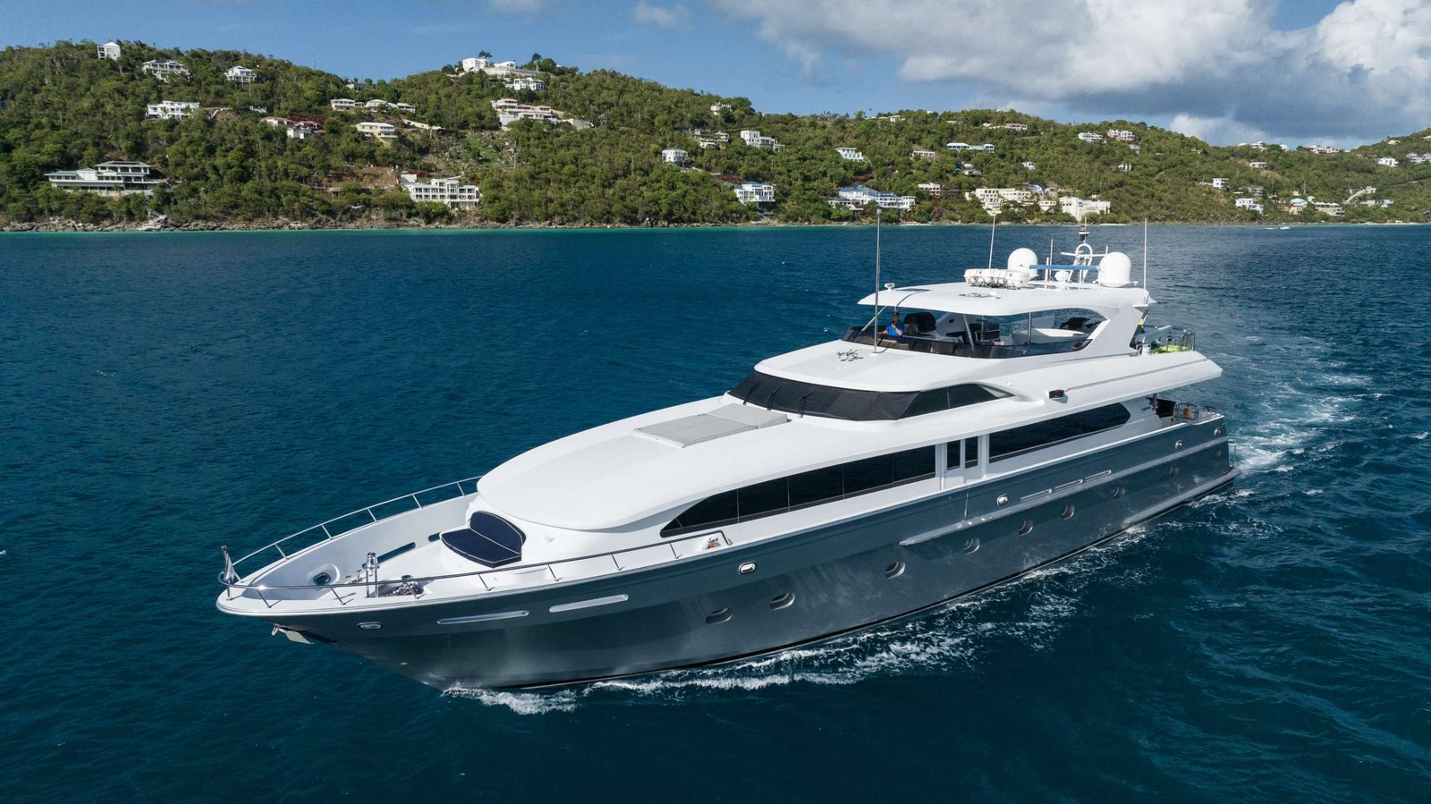 Luxury white multi-deck motor yacht cruising in turquoise sea with a green hillside and houses in the background, under a blue sky on calm waters.