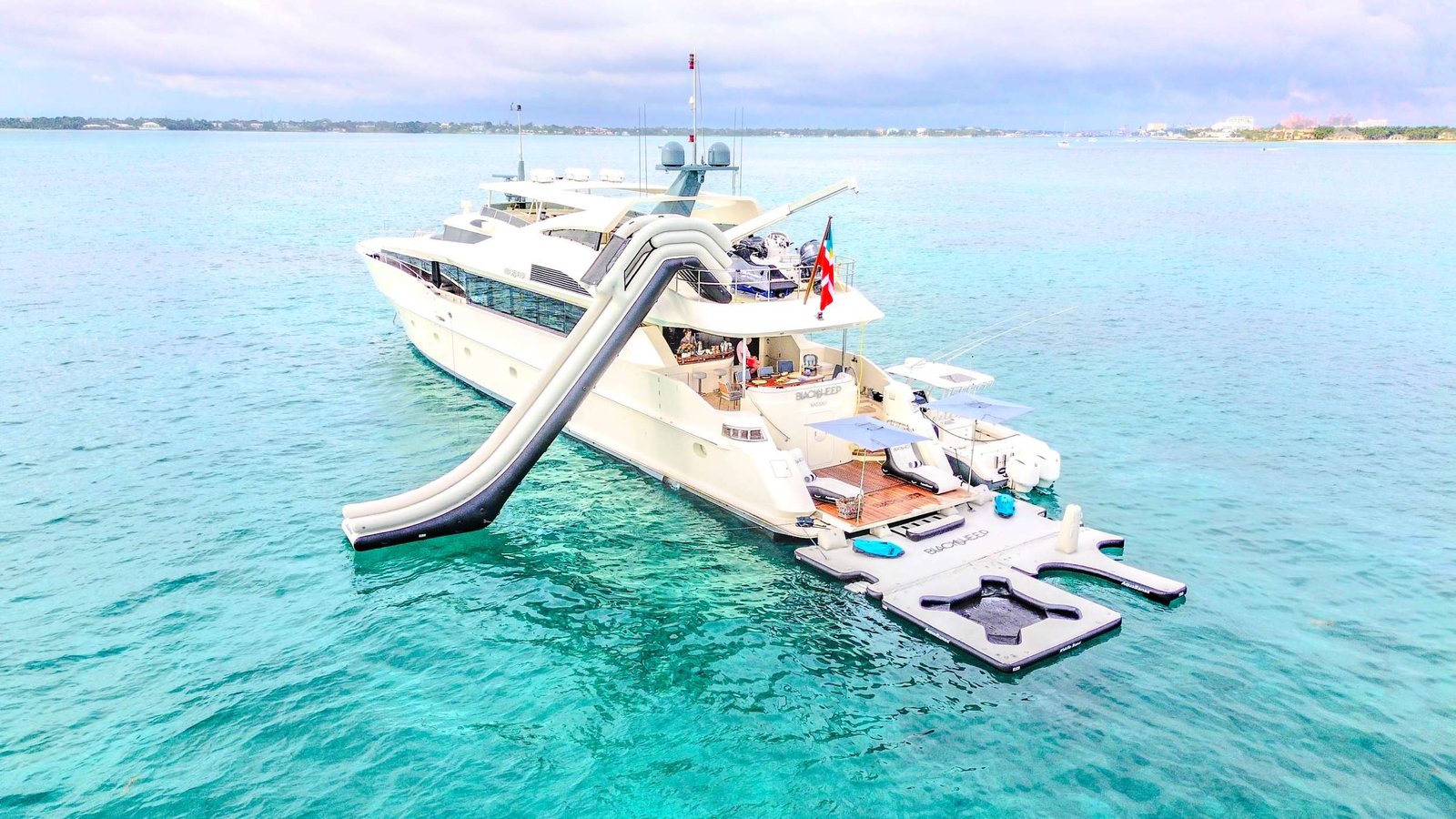 White luxury yacht anchored in turquoise waters with a floating platform nearby and distant shoreline visible.