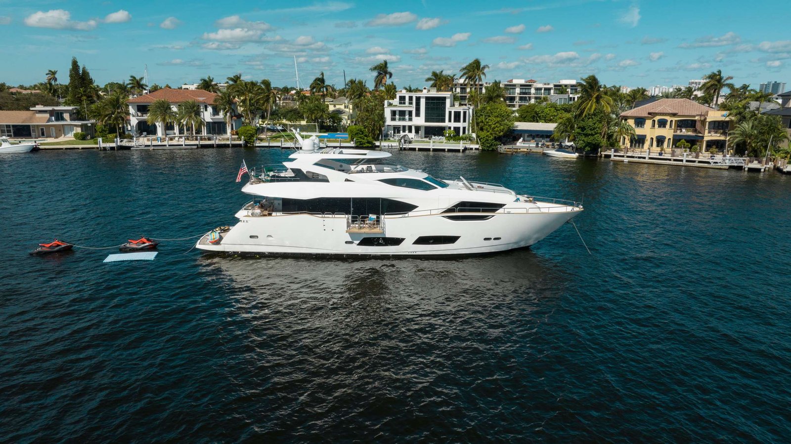 White luxury motor yacht anchored in a canal with waterfront villas and palm trees along the shore under a blue sky