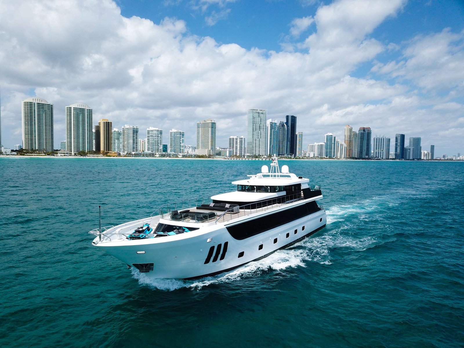 White luxury yacht sails through turquoise water with a city skyline in the distance under a blue, partly cloudy sky.