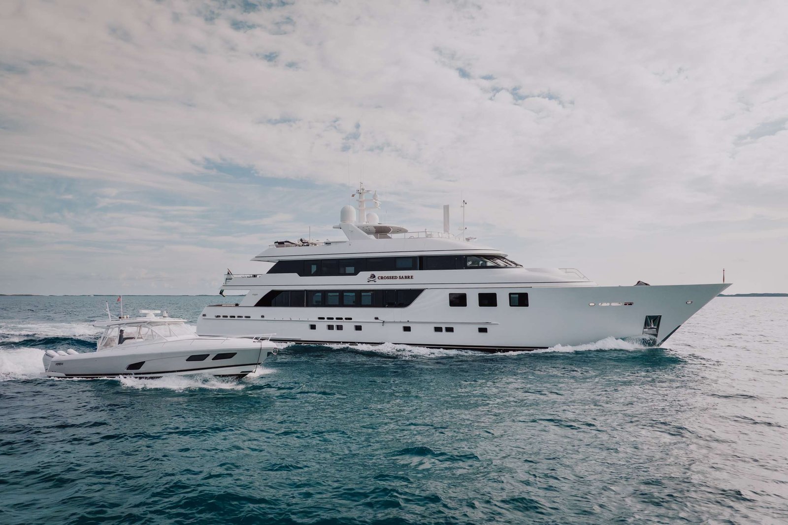 Large white luxury motor yacht gliding through blue-green water under a cloudy sky, with a smaller boat in the foreground.