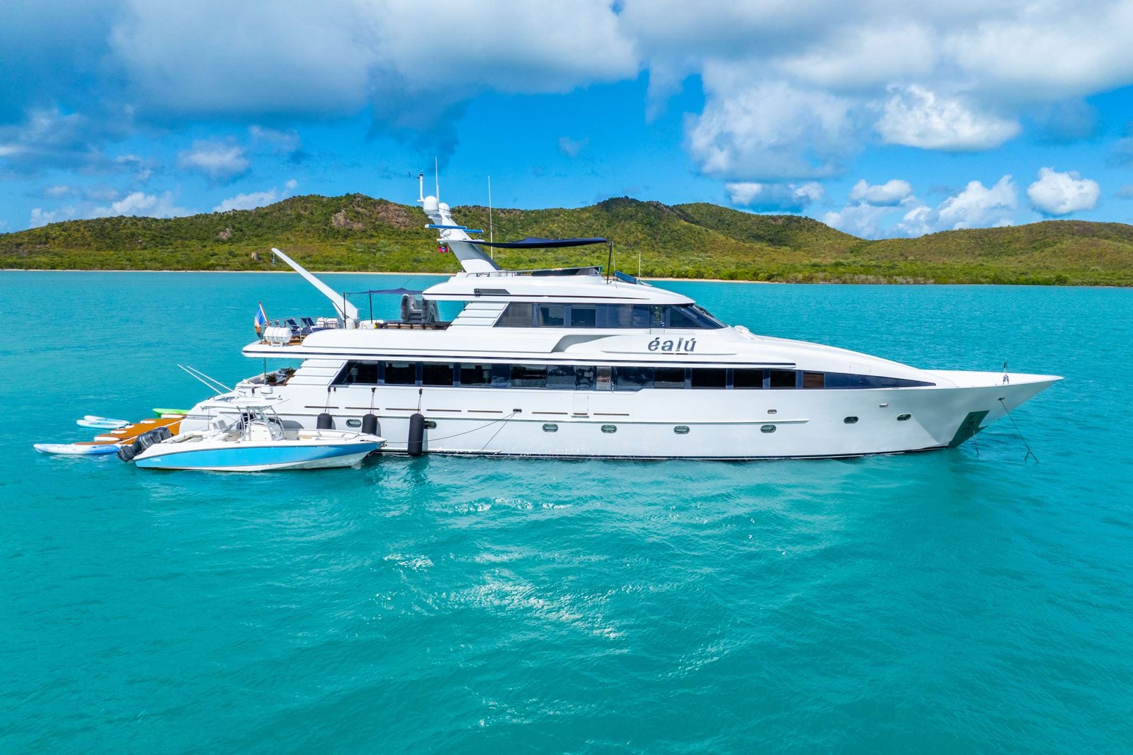 White luxury motor yacht anchored in a turquoise bay with green hills and a partly cloudy blue sky in the background, plus smaller boats nearby.