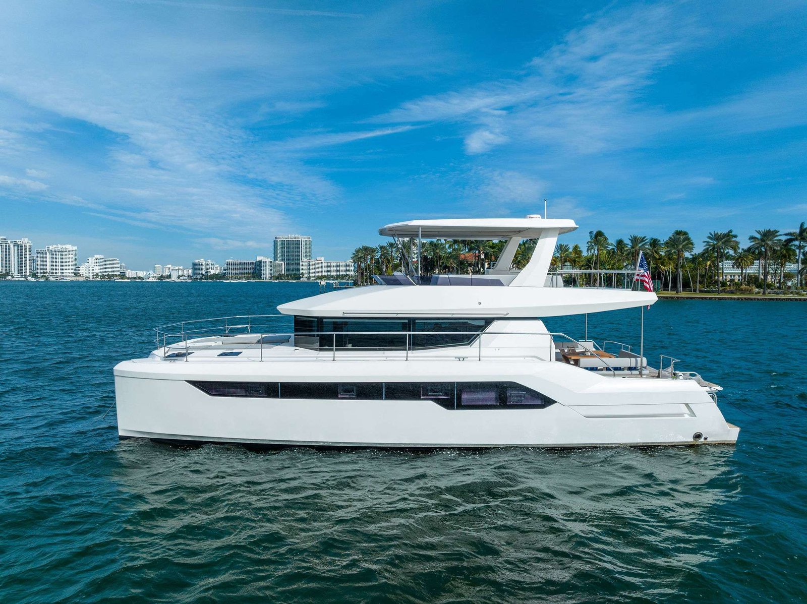 White luxury yacht on blue water with a city skyline and palm trees in the background, under a clear sky. The boat has an American flag at the stern.