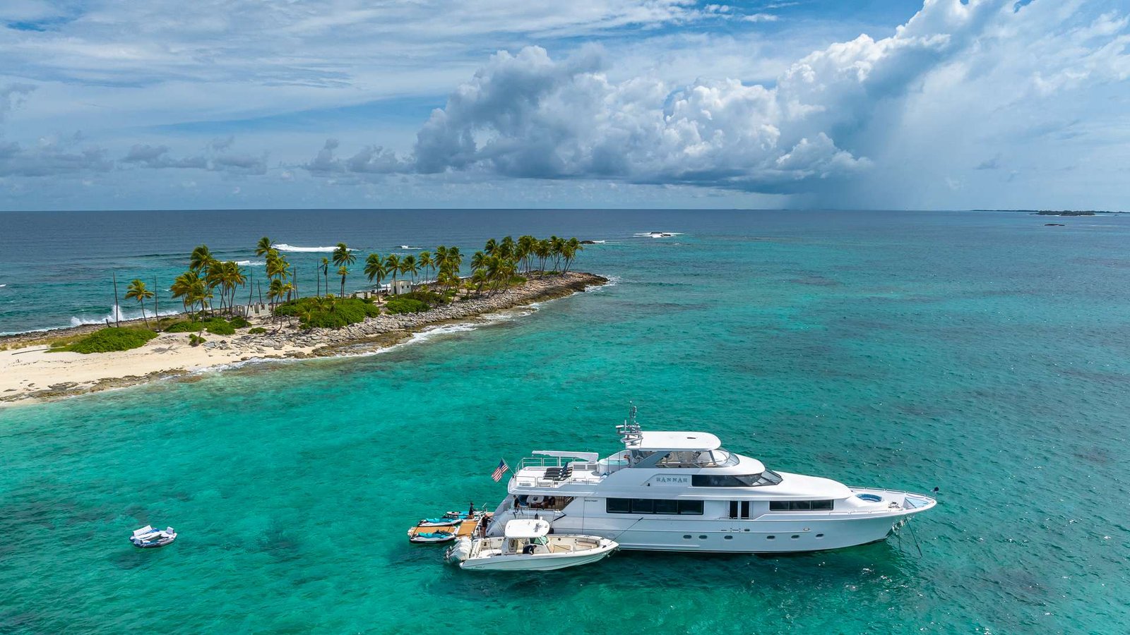 Small tropical island with palm trees and a rocky edge, turquoise water, and a large white yacht docked nearby in the foreground.