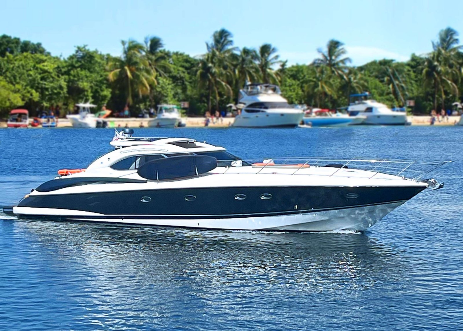 White luxury motor yacht cruising on blue water with a tropical beach and several boats anchored along a palm-lined shore in the background.