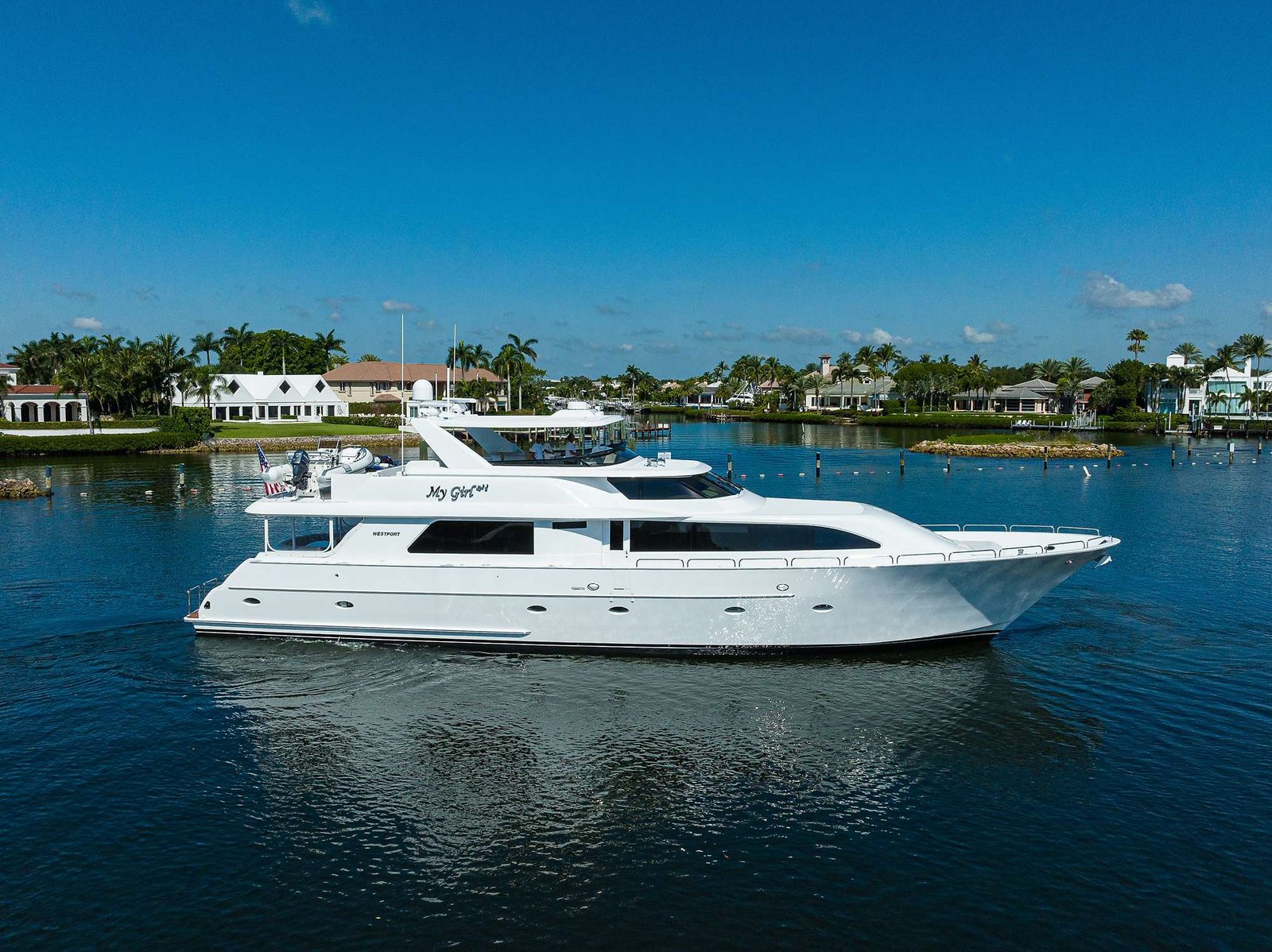 White luxury motor yacht docked in a marina with palm-lined shorefront and houses in the background.
