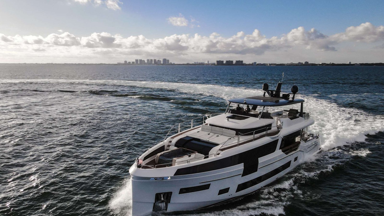 White luxury motor yacht cruising on blue water with a coastal city skyline in the distance.