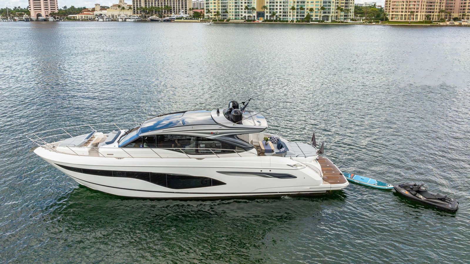 White luxury motor yacht anchored on calm water with a city waterfront and high-rise buildings in the background, including a small paddleboard nearby.