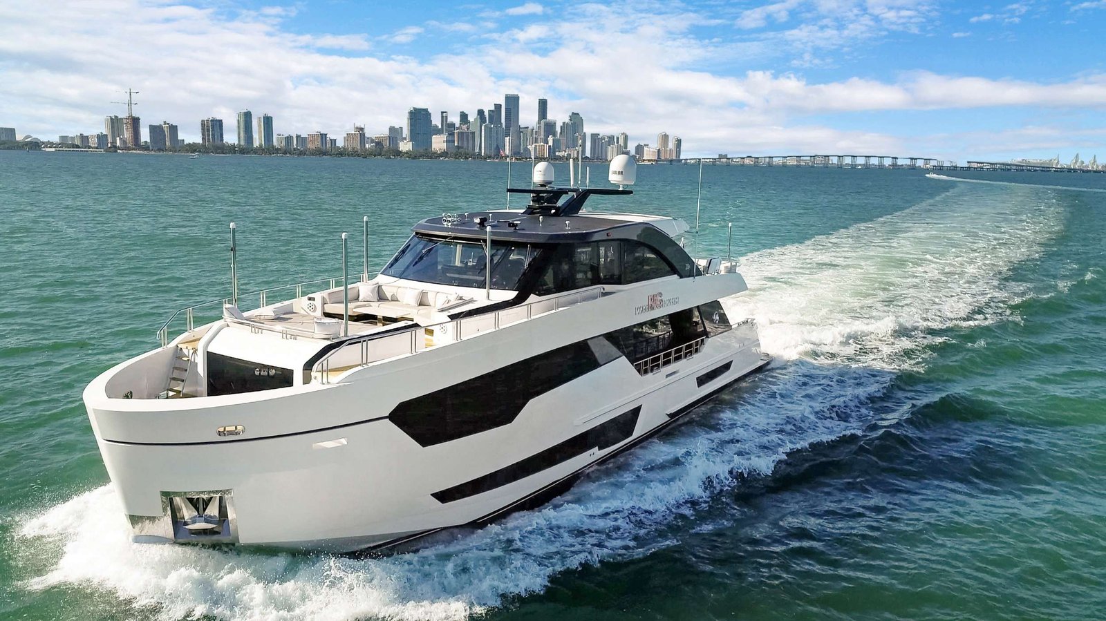 Luxurious white motor yacht speeding through turquoise water with a city skyline in the distance.