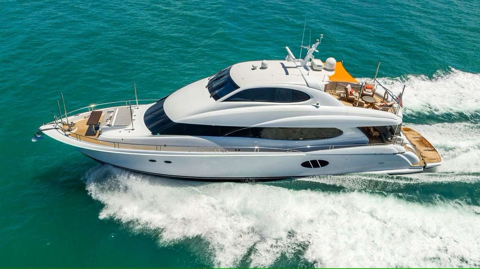 A white luxury motor yacht steams through turquoise water, leaving a frothy wake behind it. The top deck has an orange canopy.