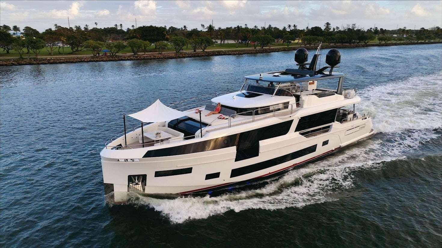 White luxury motor yacht cruising on calm water near a tree-lined shoreline, with a white canopy and sun deck on deck.