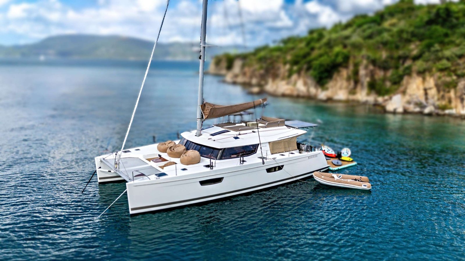 White luxury catamaran anchored in clear blue water near a rocky green coastline, with brown loungers on deck and small inflatable boats nearby.