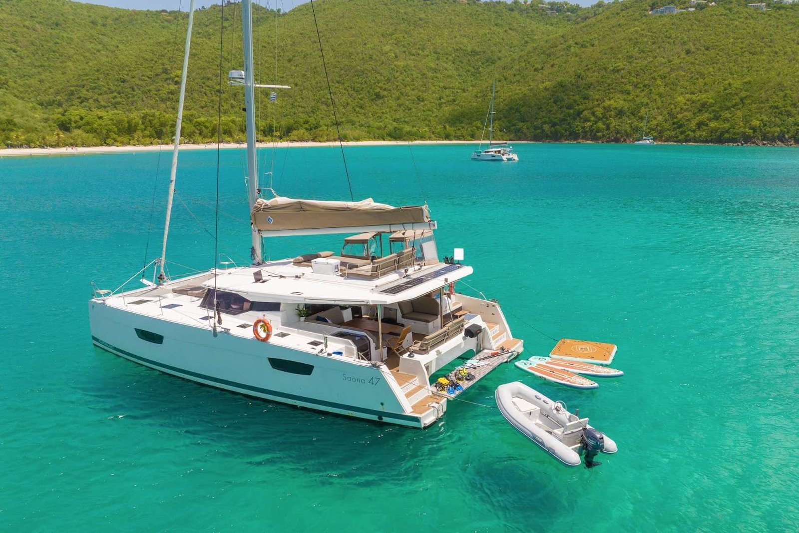 Luxurious white catamaran anchored in clear turquoise water near a sandy beach with paddle boards and a small dinghy attached to the stern