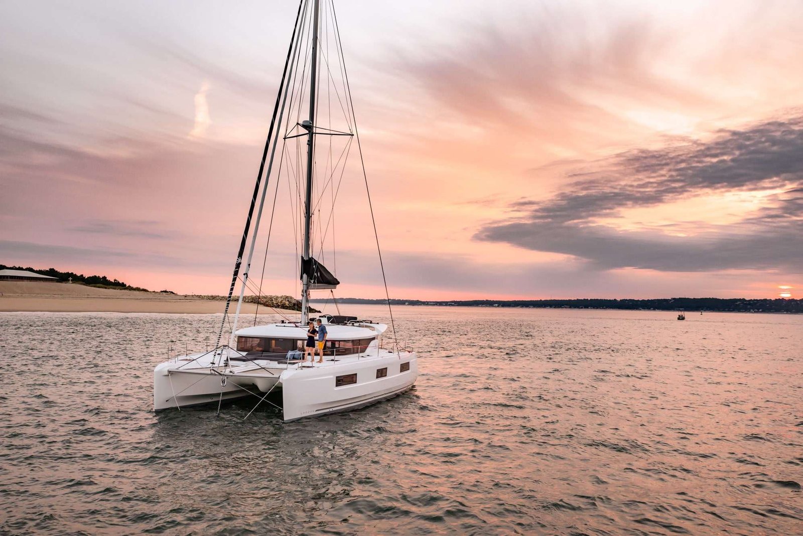 Catamaran sailboat on calm water at sunset with two people standing on the deck and colorful sky in the background
