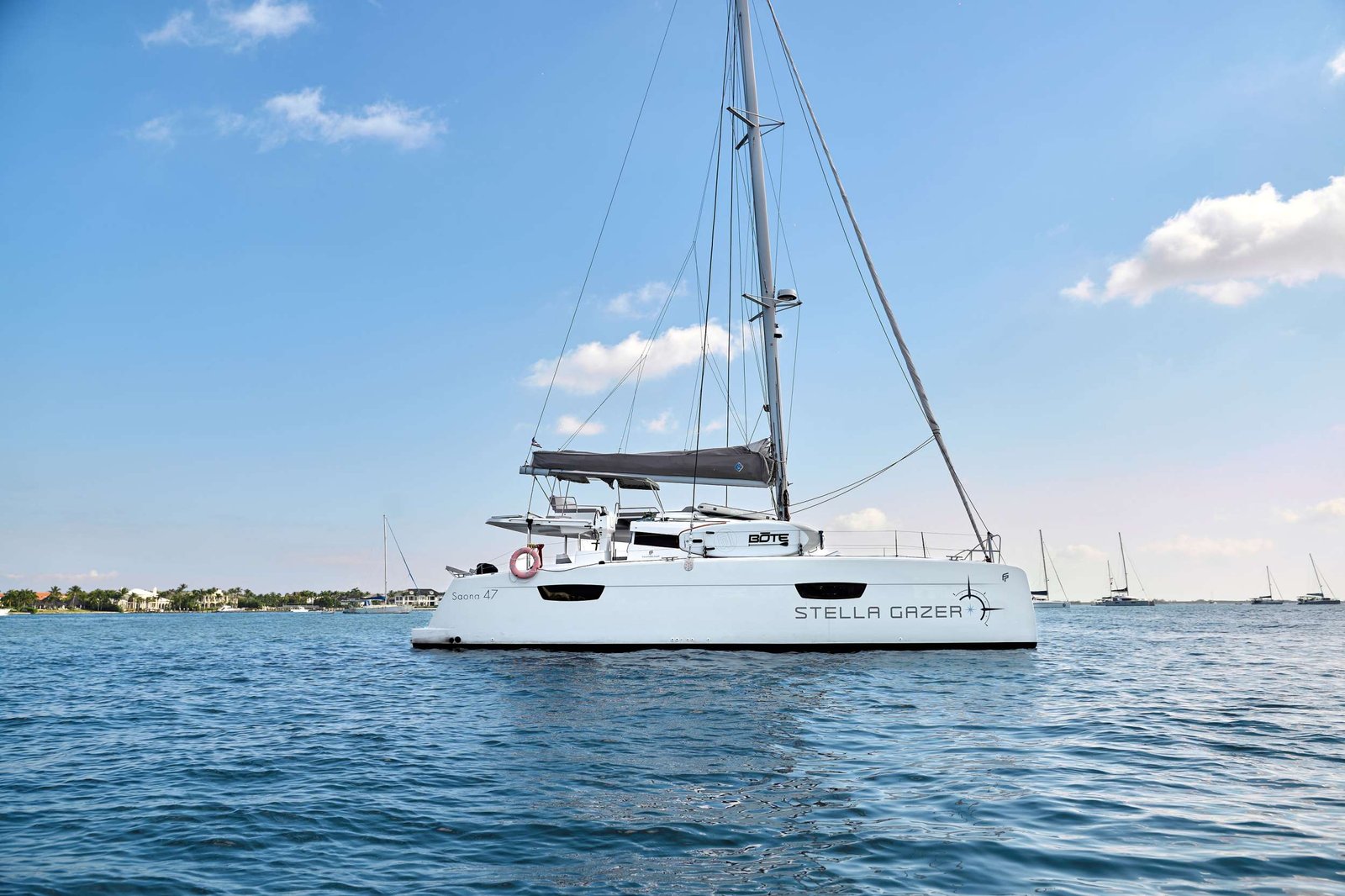 White sailing catamaran named Stella Gazer on calm blue water with a distant shoreline in view.