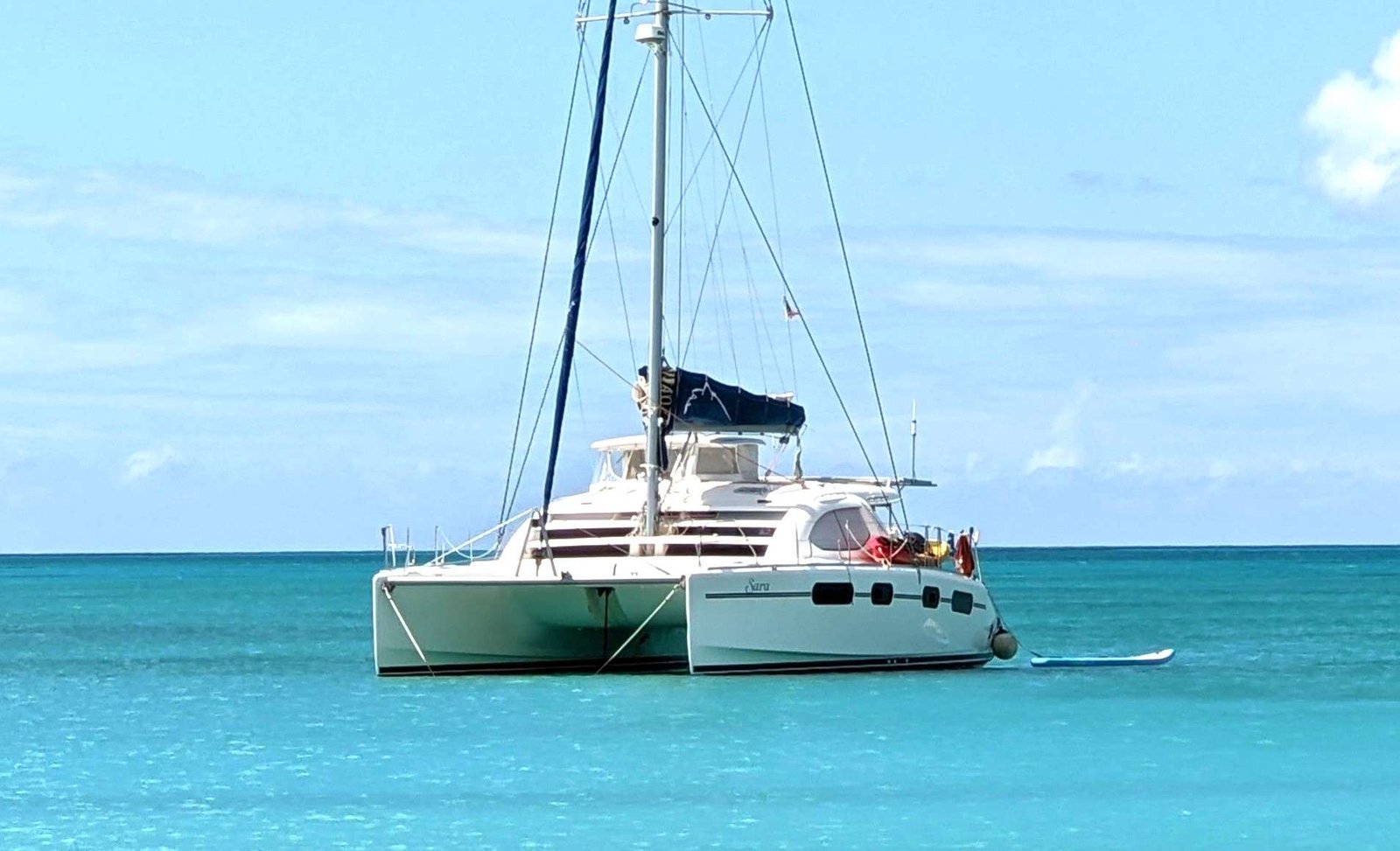 White catamaran anchored in turquoise tropical water with a paddleboard nearby and a blue sky above.
